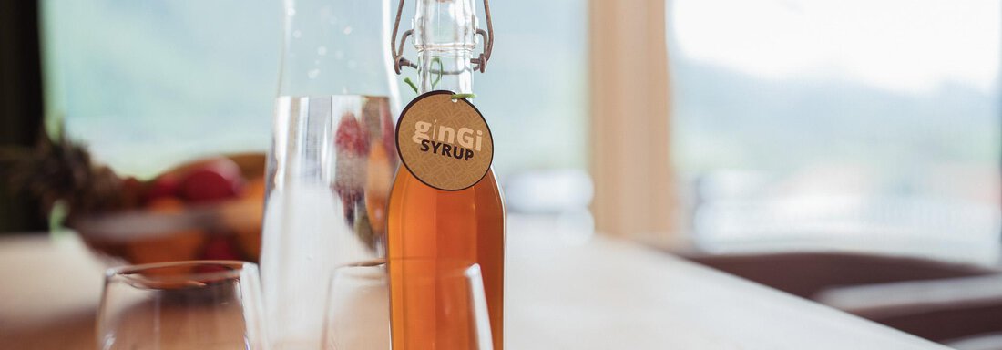 A bottle of RIng Syrup sits on a wooden table, next to it are two glasses. In the background, fresh fruit and a bright, peaceful setting can be seen. - All-Suite Resort Ötztal - Oetz