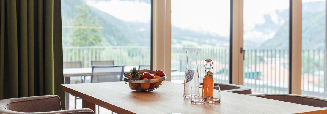 A stylish room with a table on which a bowl of fruit is placed. The windows offer a view of the mountains and plenty of natural light. - All-Suite Resort Ötztal - Oetz