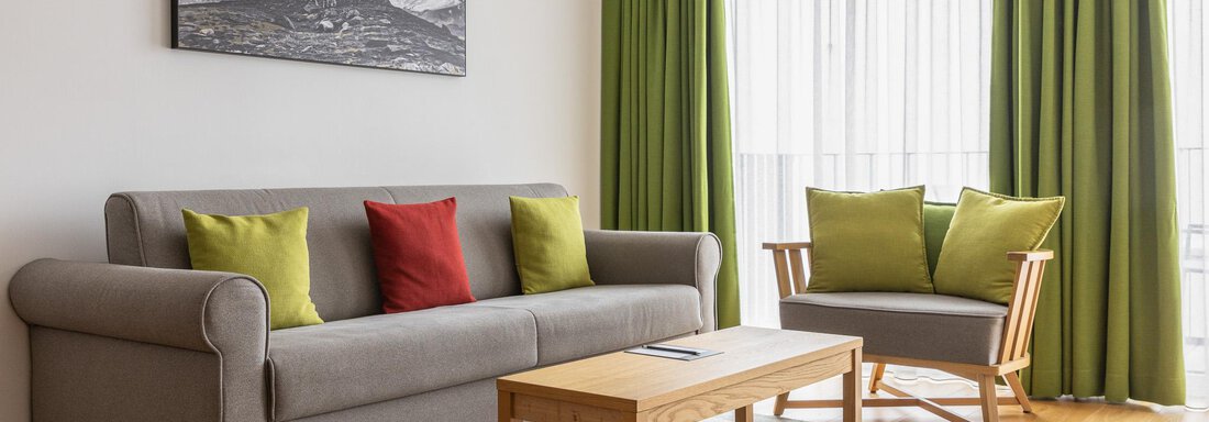 A modern living room with a gray sofa and colorful cushions. Large windows with green curtains let in plenty of light. - All-Suite Resort Ötztal - Oetz