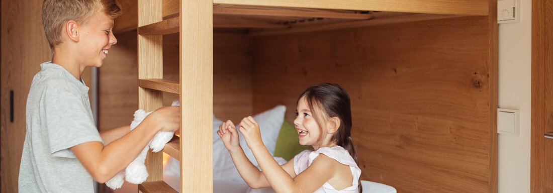 A boy is standing next to a bunk bed and smiling while a girl is sitting on the lower bed. The room is bright and inviting, with wooden paneling and white bed linens. - All-Suite Resort Ötztal - Oetz