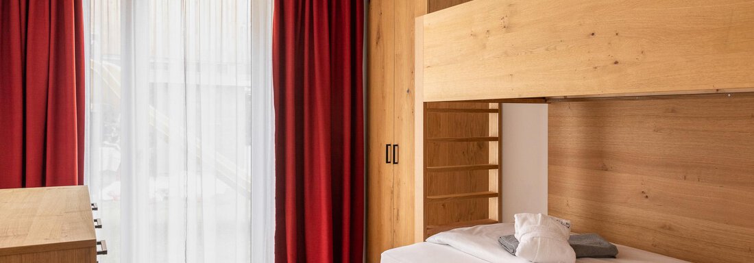 A modern bedroom with wooden furniture and red curtains. The room has large windows that let in a lot of daylight. - All-Suite Resort Ötztal - Oetz