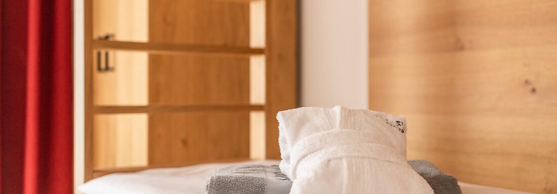 A cozy bed with fresh towels. In the background, there is a wooden wardrobe and a red curtain. - All-Suite Resort Ötztal - Oetz