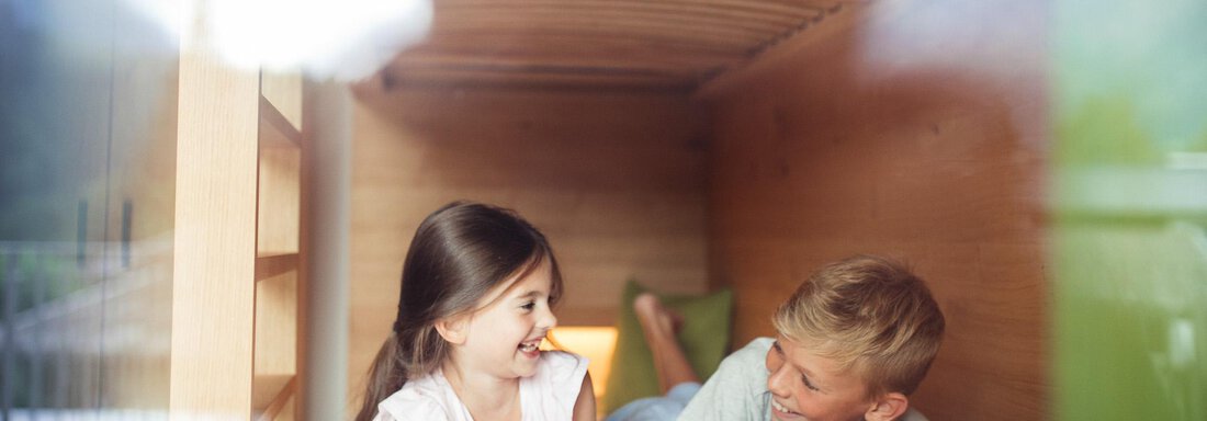 Two children are lying in a bed and laughing with each other. The cozy wooden surroundings create a relaxed atmosphere. - All-Suite Resort Ötztal - Oetz