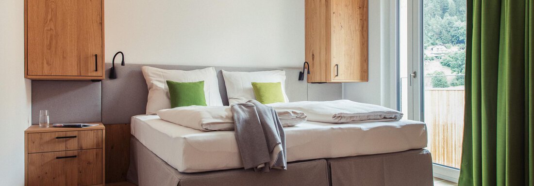 A modern bedroom with a large bed and wooden furniture.  
Large windows with green curtains provide plenty of daylight. - All-Suite Resort Ötztal - Oetz