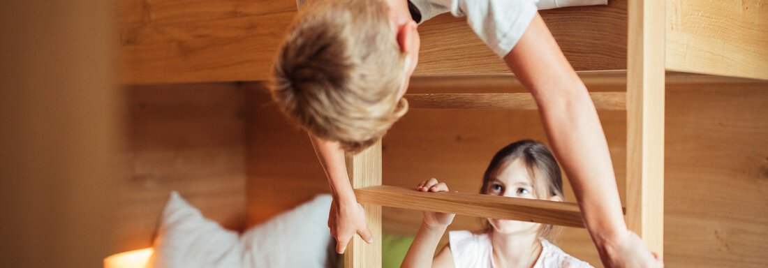 Two children are playing in a bunk bed. A boy is leaning over the ladder and looking at a girl who is sitting below. - All-Suite Resort Ötztal - Oetz