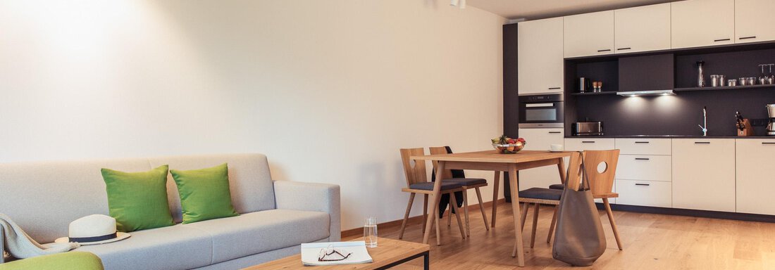 A modern living room with a gray sofa and a wooden table. The kitchen is done in neutral colors and is well equipped. - All-Suite Resort Ötztal - Oetz