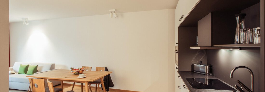 A modern living room with a dining table and chairs. The kitchen is open and well-equipped, with wooden flooring and bright light. - All-Suite Resort Ötztal - Oetz