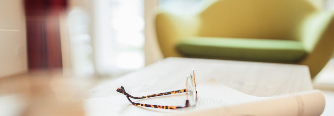A pair of glasses is lying on a table next to a rolled-up sheet of paper. In the background, a green sofa can be seen. - All-Suite Resort Ötztal - Oetz