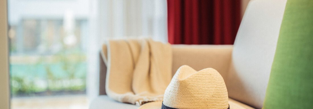 A cozy sofa with a straw hat and a blanket. In the background, light and colors of a modern interior can be seen. - All-Suite Resort Ötztal - Oetz