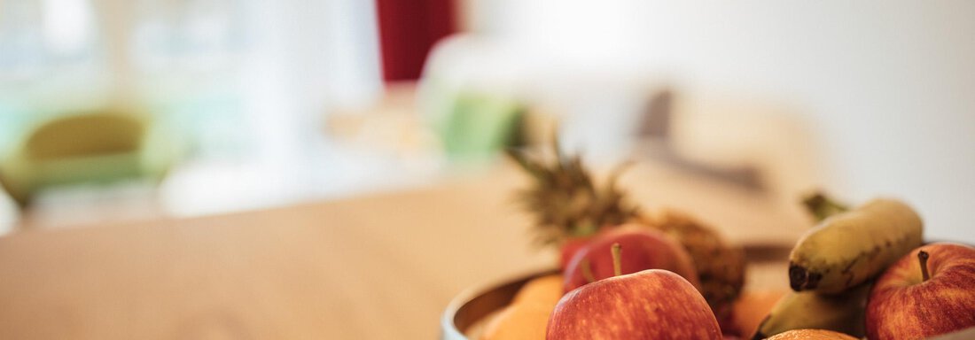 A bowl of fresh fruit, including apples, oranges, and a banana. In the background, a bright room with a cozy armchair can be seen. - All-Suite Resort Ötztal - Oetz