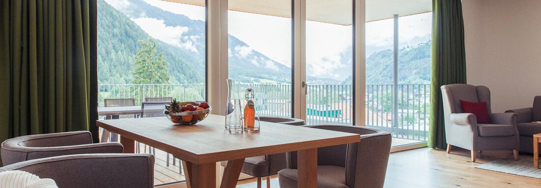 A modern living room with large windows that provide a view of the mountains. The space is bright and friendly with a dining table and cozy armchairs. - All-Suite Resort Ötztal - Oetz