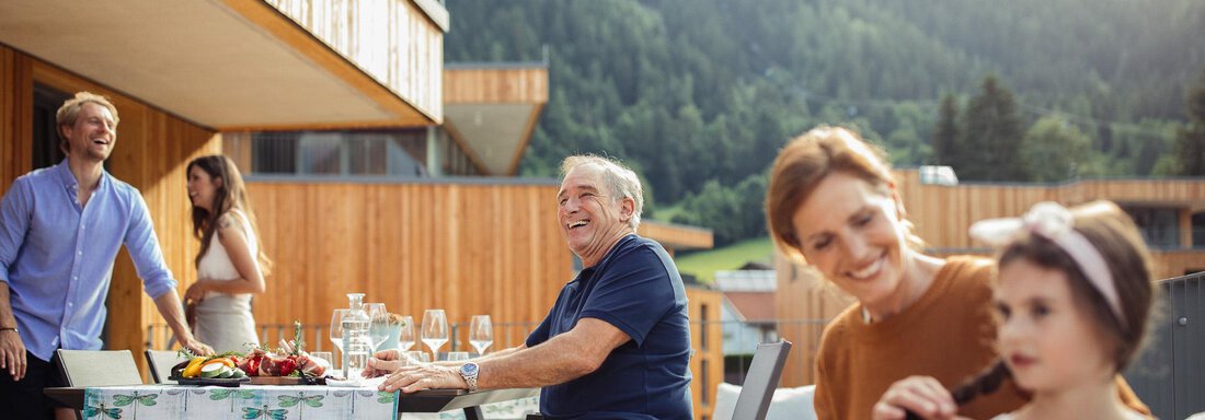 A group of people sits relaxed on a terrace with a view of the mountains. There is a table with a selection of foods and drinks. - All-Suite Resort Ötztal - Oetz