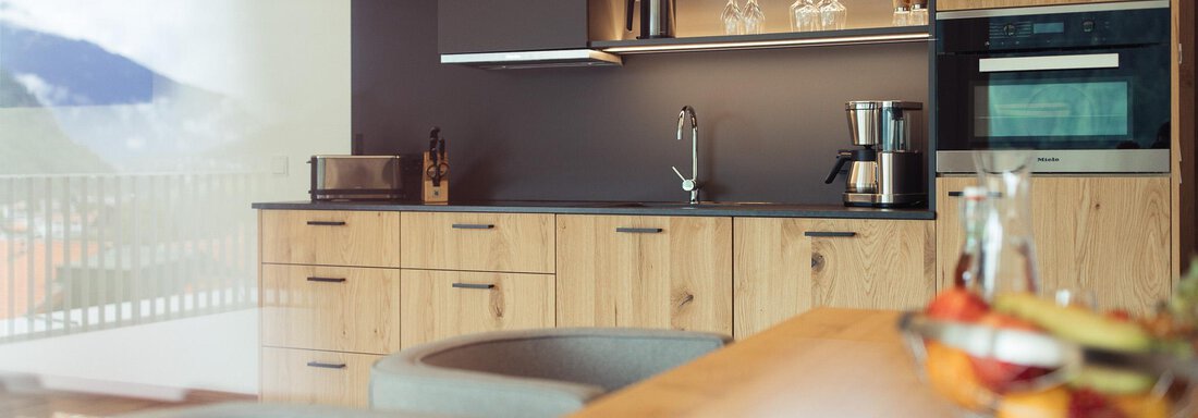 Modern kitchen with wooden cabinets and elegant appliances. A table with a fruit bowl is visible in the foreground. - All-Suite Resort Ötztal - Oetz
