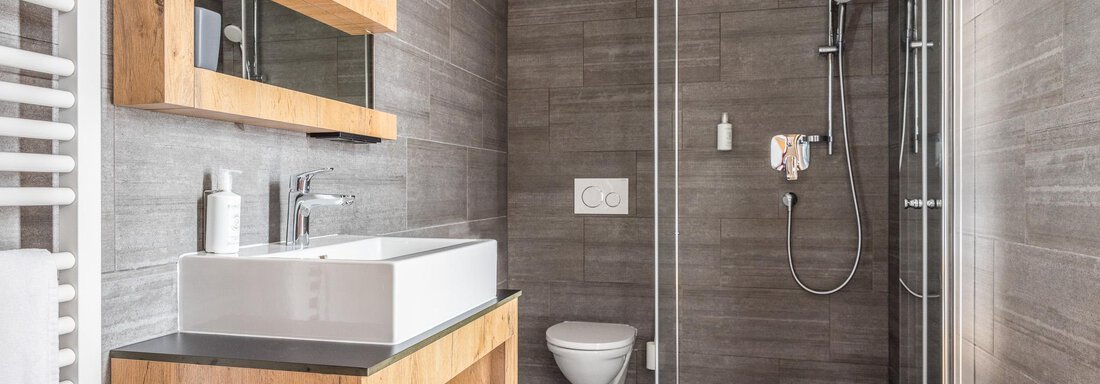 A modern bathroom with wood details and gray tiles. It features a shower, a sink, and a stylish mirror dresser. - All-Suite Resort Ötztal - Oetz