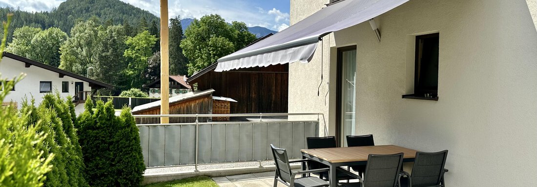 Eine gemütliche Terrasse mit einem Esstisch und Stühlen, umgeben von grünen Pflanzen. Im Hintergrund sind Hügel und ein blauer Himmel sichtbar. - ALPsmart - Oetz