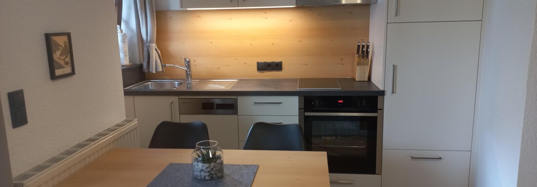 Wohnküche Eine moderne Küche mit hellen Holz- und weißen Möbeln. Der Tisch ist schlicht gedeckt und es gibt eine große Fensterfläche für viel Tageslicht. - Apart Swoboda - Oetz