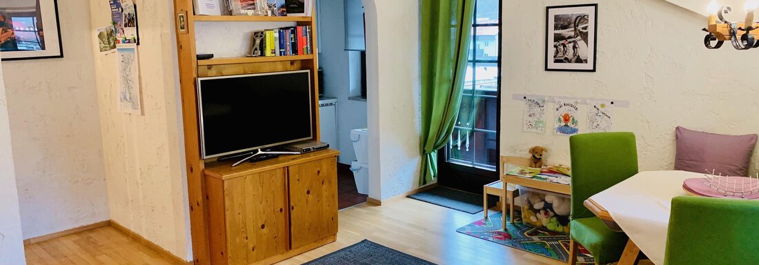 A cozy living room with wooden shelves and a television. The room is bright and cheerfully designed, featuring a small dining area and green chairs. - Appartement Azalea - Oetz