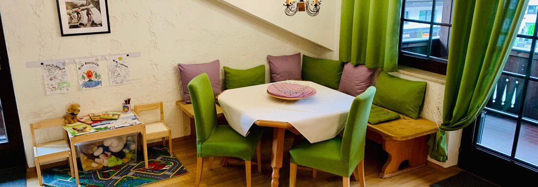 A cozy dining area with a round table and green chairs. The window is adorned with green curtains, and there is a small play area for children. - Appartement Azalea - Oetz