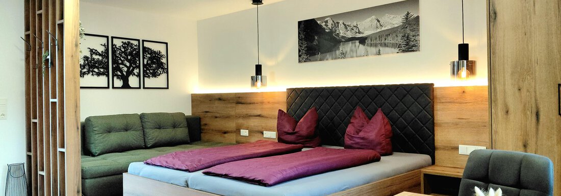 A modern bedroom with a large wooden bed and purple pillows. In the background, there is a cozy living area with a gray couch and a table. - Haus Gstrein - Oetz