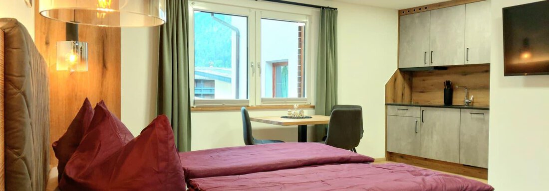 A cozy room with a double bed and red pillows. The room has a small kitchenette and plenty of daylight through the windows. - Haus Gstrein - Oetz