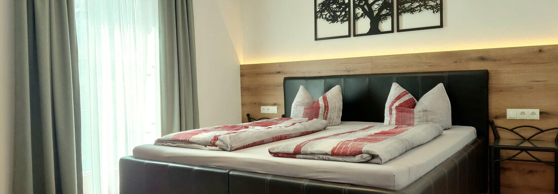 A modern bedroom with a large double bed and decorative pillows. The windows are dressed with curtains and there are artworks on the wall. - Haus Gstrein - Oetz