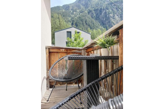 A cozy balcony with a round chair and a table. In the background, green mountains and a modern building can be seen. - Haus Gstrein - Oetz