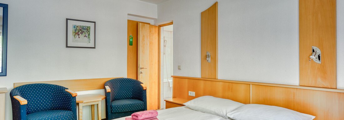 A bright room with a double bed and two blue armchairs. On the bed lie two pink towels. - Seehüter´s Hotel Seerose - Oetz