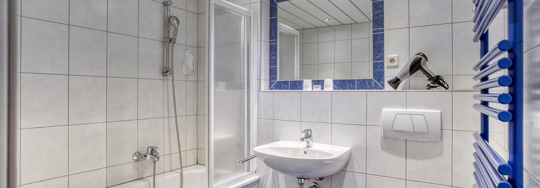 A modern bathroom with a bathtub, shower, and a sink. The walls are light tiled and there is a large mirror. - Seehüter´s Hotel Seerose - Oetz