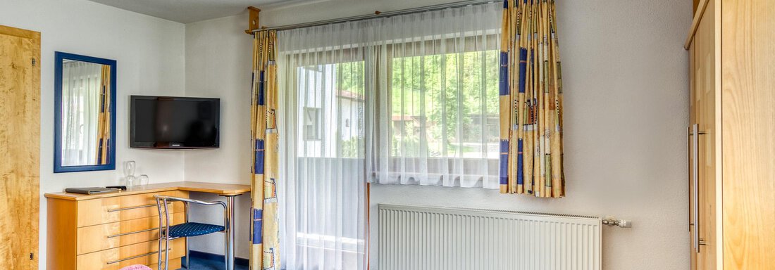 A bright room with two beds, a television, and a desk. The curtains are colorful, and there are two towels on the beds. - Seehüter´s Hotel Seerose - Oetz