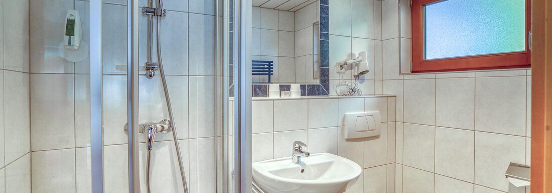 A modern bathroom with a shower, a sink, and a toilet. The walls are tiled, and there is a window for natural light. - Seehüter´s Hotel Seerose - Oetz