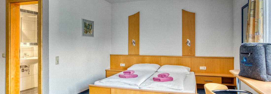A simple room with a double bed and blue carpet.  
Next to the bed are two towels and there is a chair as well as access to the bathroom. - Seehüter´s Hotel Seerose - Oetz