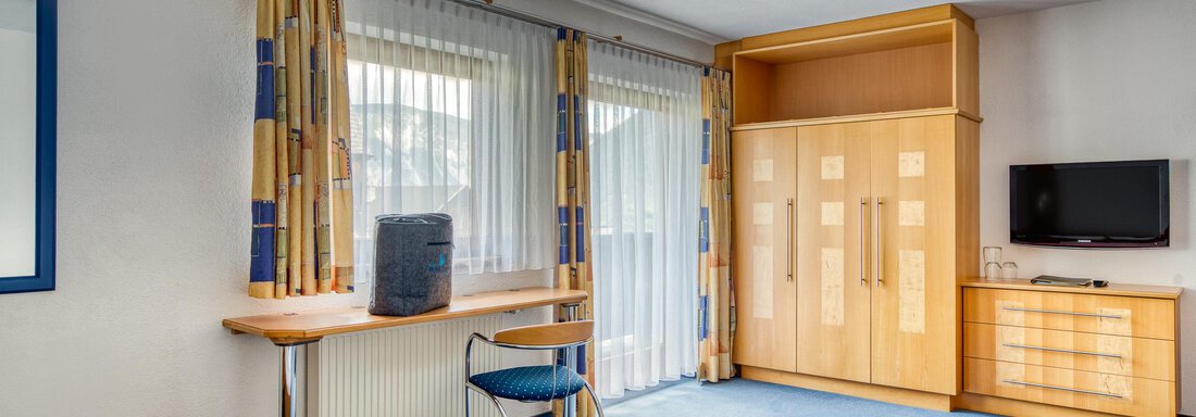 A bright room with large windows and simple, modern furnishings. There is a desk, a chair, and a television. - Seehüter´s Hotel Seerose - Oetz