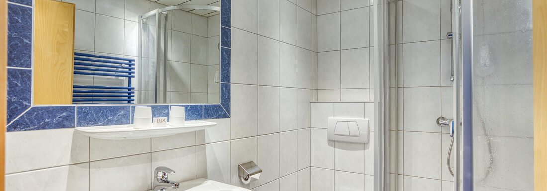 A modern bathroom with a shower, sink, and toilet. The walls are tiled in light colors, and a mirror reflects the space. - Seehüter´s Hotel Seerose - Oetz