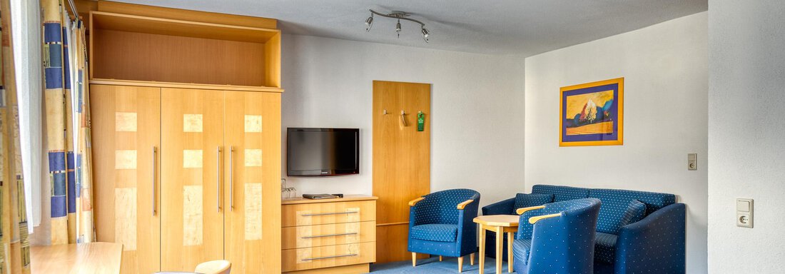 A cozy hotel room with blue furniture and a desk. There is a television and plenty of space to relax. - Seehüter´s Hotel Seerose - Oetz