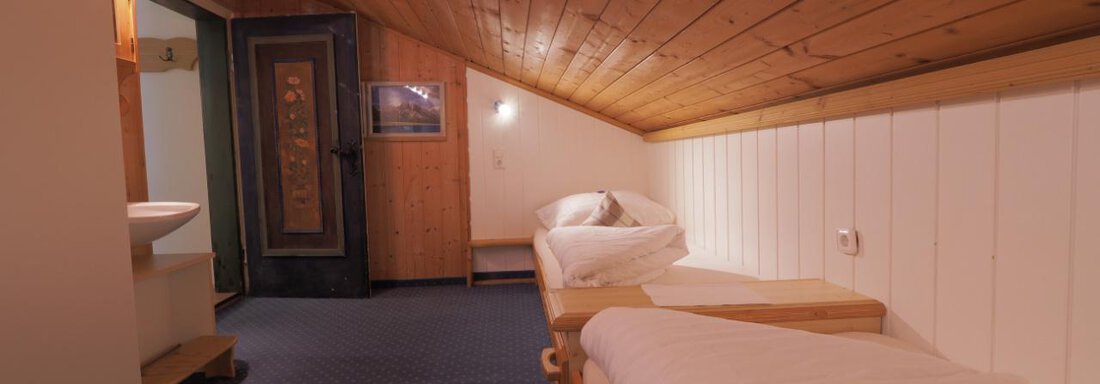 Ein gemütlicher Dachraum mit zwei Einzelbetten und einem Holzdecken. Die Wände sind hell und der Boden hat einen blauen Teppich. - bei Herrmann´s - Sautens