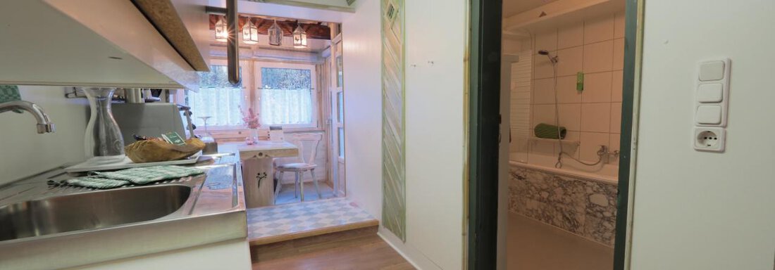 Eine helle Küche mit Holzregalen und einem angrenzenden Essbereich. Im Hintergrund ist ein Badezimmer mit einer modernen Badewanne zu sehen. - bei Herrmann´s - Sautens