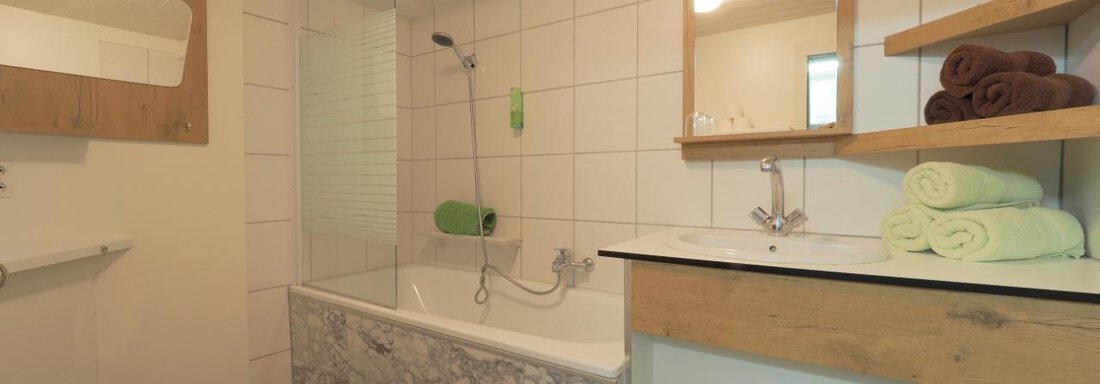Ein modernes Badezimmer mit einer Badewanne und einer Dusche. Es gibt Holz- und Fliesenakzente sowie Handtücher in verschiedenen Farben. - bei Herrmann´s - Sautens