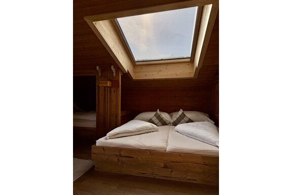 Ein gemütliches Wohn- Schlafzimmer mit einem Doppelbett und Holzdecken. Ein Dachfenster lässt Tageslicht herein und ist mit einem UV Rollo gegen Sonneneinstrahlung und Wärme geschützt. - bei Herrmann´s - Sautens