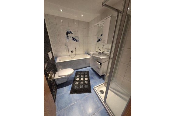 A modern bathroom with a shower, bathtub, and sink. The floor is tiled in blue and a black carpet lies in front of the sink. - Ferienhof Strigl - Sautens
