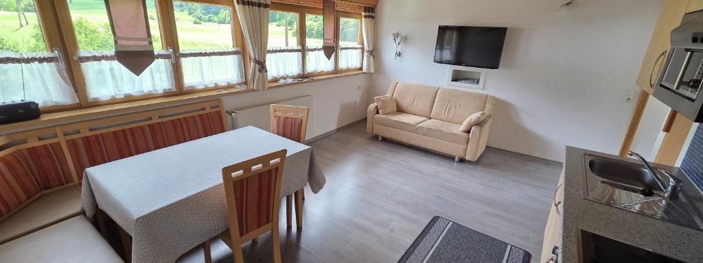 A bright, modern living and dining room with wooden ceilings and large windows. There is a dining table, a couch, and a kitchenette. - Ferienhof Strigl - Sautens