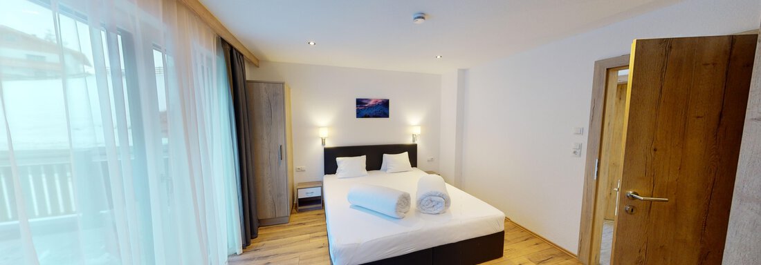 A modern bedroom with a double bed and large windows. The room is cheerful and bright, with wooden flooring and simple furniture. - A CASA "Rubin Chalet & Studio Resort" - Sölden