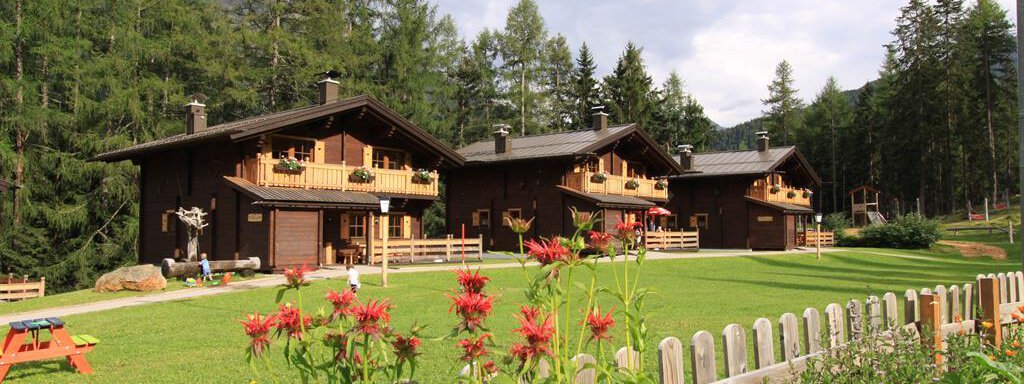 Three idyllic wooden houses surrounded by green meadows and trees. In front, colorful flowers bloom in a well-kept garden. - Almdorf Waldesruh - Sölden