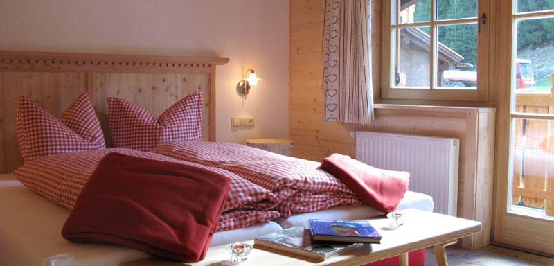 A cozy bedroom with a large bed and wooden decor. The windows offer a view of nature outside. - Almdorf Waldesruh - Sölden