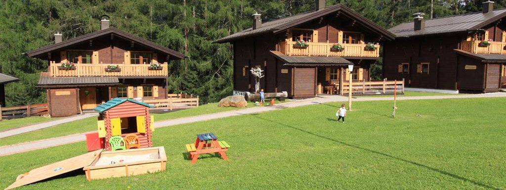 Beautiful wooden houses in a green landscape with mountains in the background. A playground with a small hut and play equipment is visible in the foreground. - Almdorf Waldesruh - Sölden
