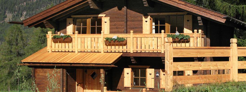 A cozy wooden house with a balcony and green planters. It is located in a rural setting with mountains in the background. - Almdorf Waldesruh - Sölden