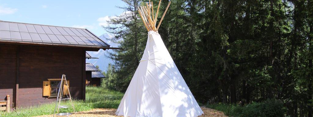 A tipi stands in a light forest environment. In the background, a wooden building can be seen. - Almdorf Waldesruh - Sölden