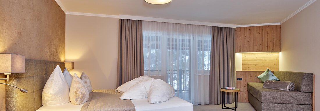 Familienzimmer A cozy hotel room with a large double bed and a seating area. The room has windows with curtains and a warm, inviting color scheme. - Alp Resort Tiroler Adler - Sölden