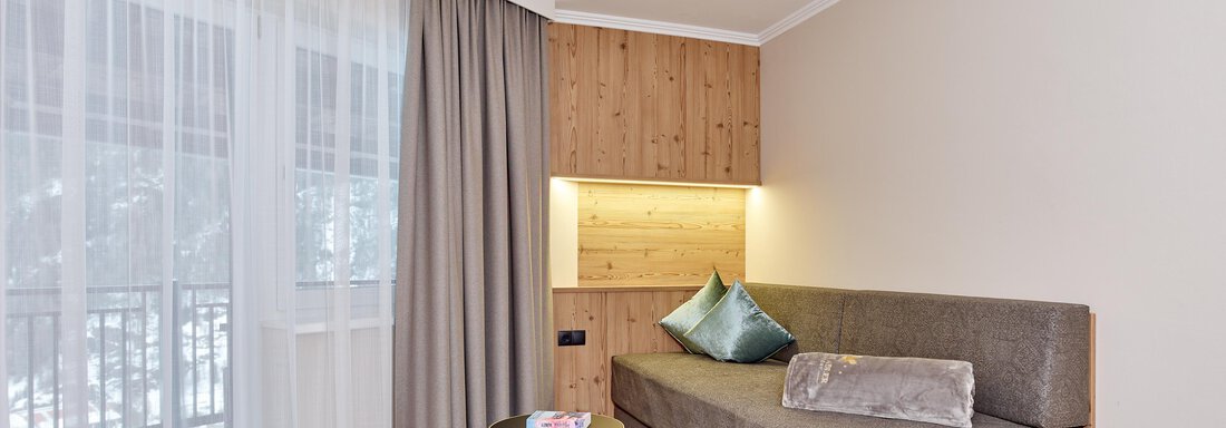 A modern living room with a gray sofa and a small table. Large windows with curtains let in plenty of light. - Alp Resort Tiroler Adler - Sölden