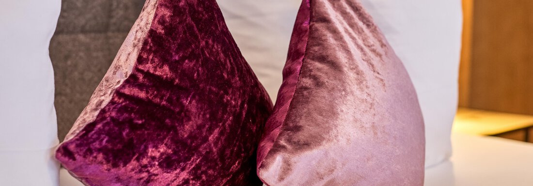 Two decorative pillows in shades of purple and pink are placed on a bed. The background features white pillows and a warm, cozy atmosphere. - Alp Resort Tiroler Adler - Sölden