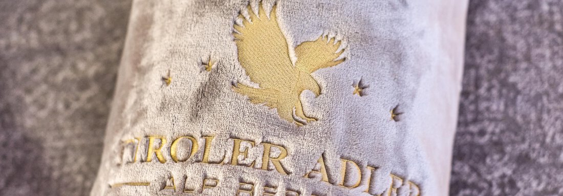 An elegant cushion with the logo of the Tyrolean Eagle Alp Resorts. The design is in gold on a velvety background. - Alp Resort Tiroler Adler - Sölden